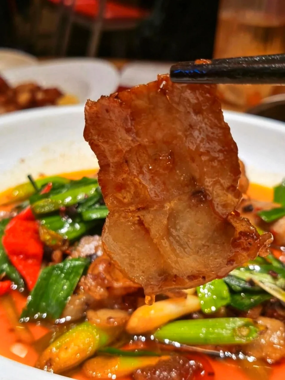 Slice of twice-cooked pork lifted by chopsticks over leeks and chili